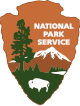 The National Park Service logo shaped like an arrowhead, featuring a tree, mountain, lake, and bison, with "National Park Service" written in white text.