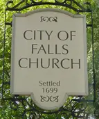 A white sign with black text reads "City of Falls Church. Settled 1699." The sign is bordered with decorative metal and is surrounded by green foliage in the background.